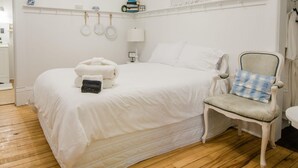 Une chambre à coucher blanche, avec un lit, une chaise et une étagère ornée d’objets décoratifs.