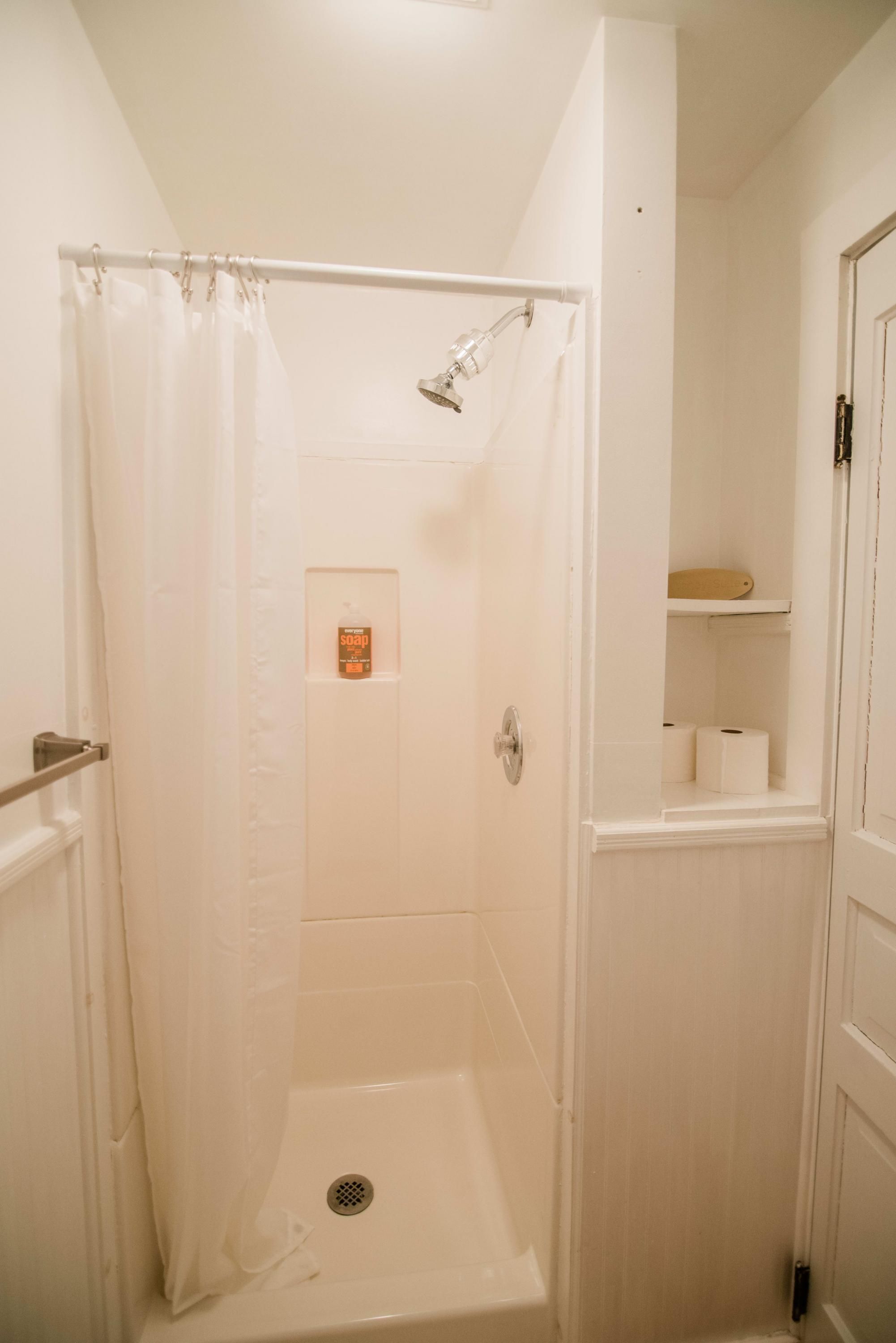Un baño pequeño y blanco con ducha, inodoro y un estante con artículos de aseo.