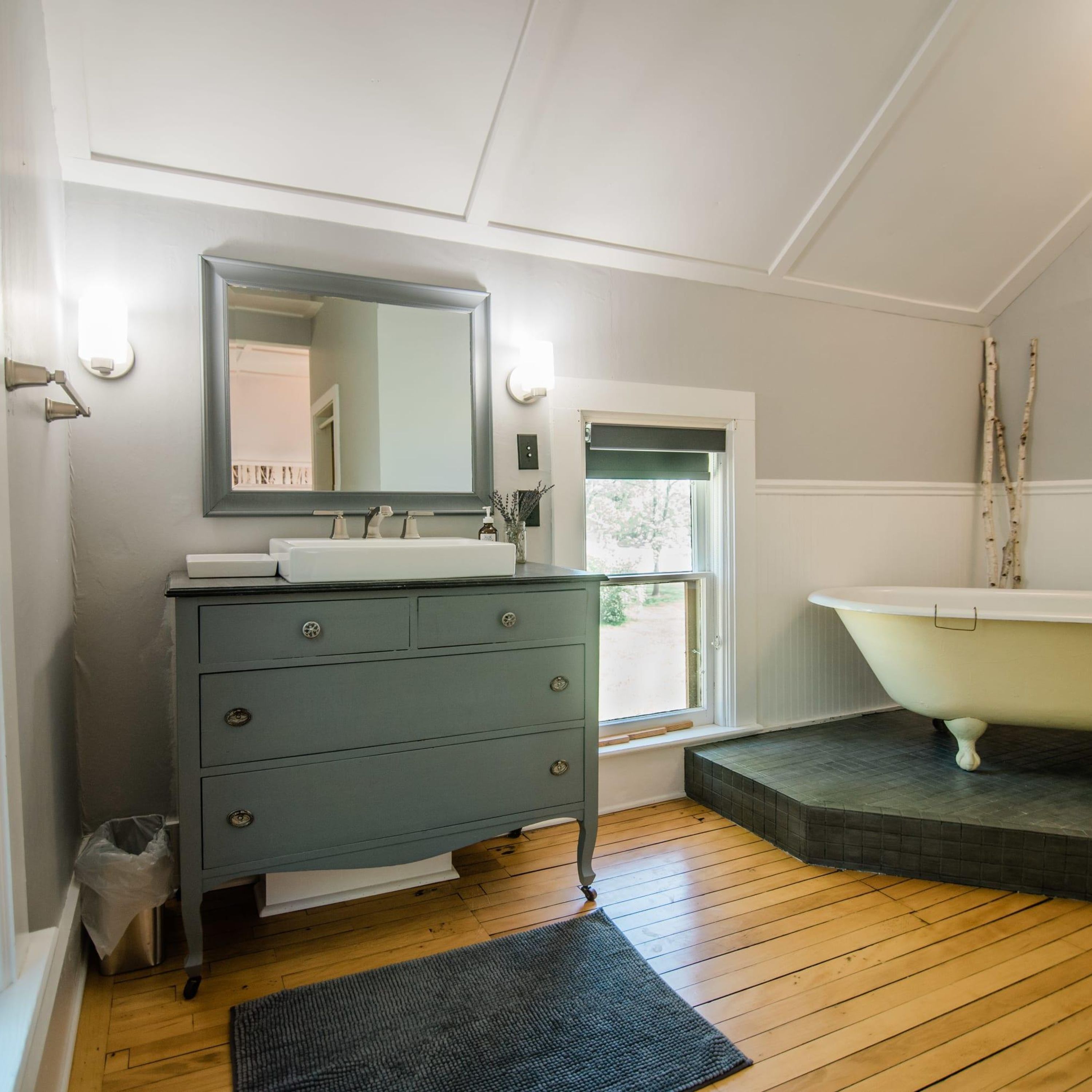 A bathroom with a grey vanity, a white sink, a large mirror, a white bathtub, and a window with a view of greenery.