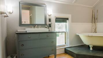 A bathroom with a grey vanity, a white sink, a large mirror, a white bathtub, and a window with a view of greenery.