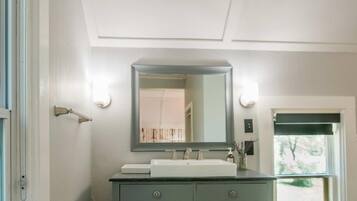 Baño con lavamanos blanco, un espejo y una lámpara sobre él. Se ve una ventana con vistas a árboles.