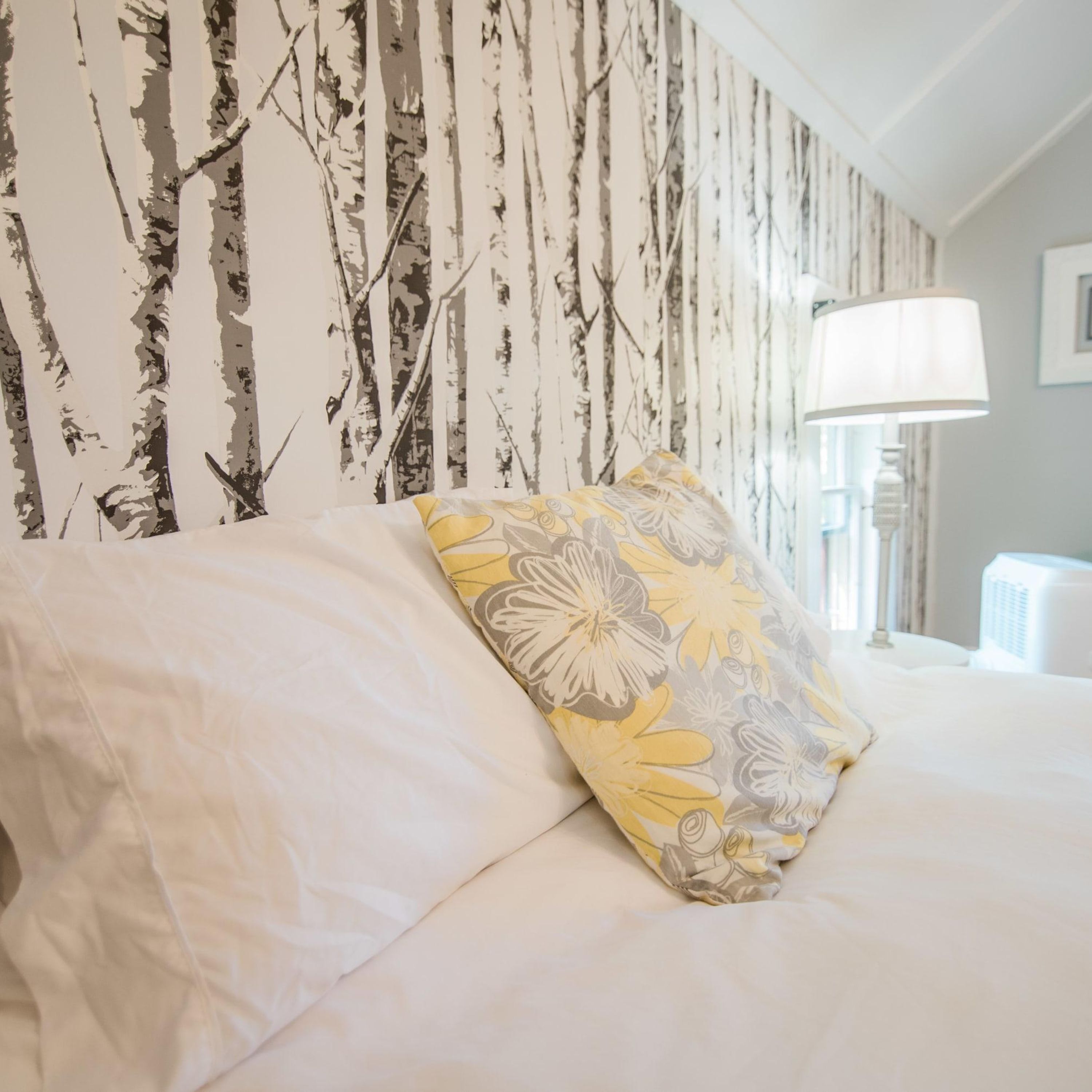 A neatly made bed with a floral pillow and a plain pillow, set against a wall with a birch tree pattern wallpaper.
