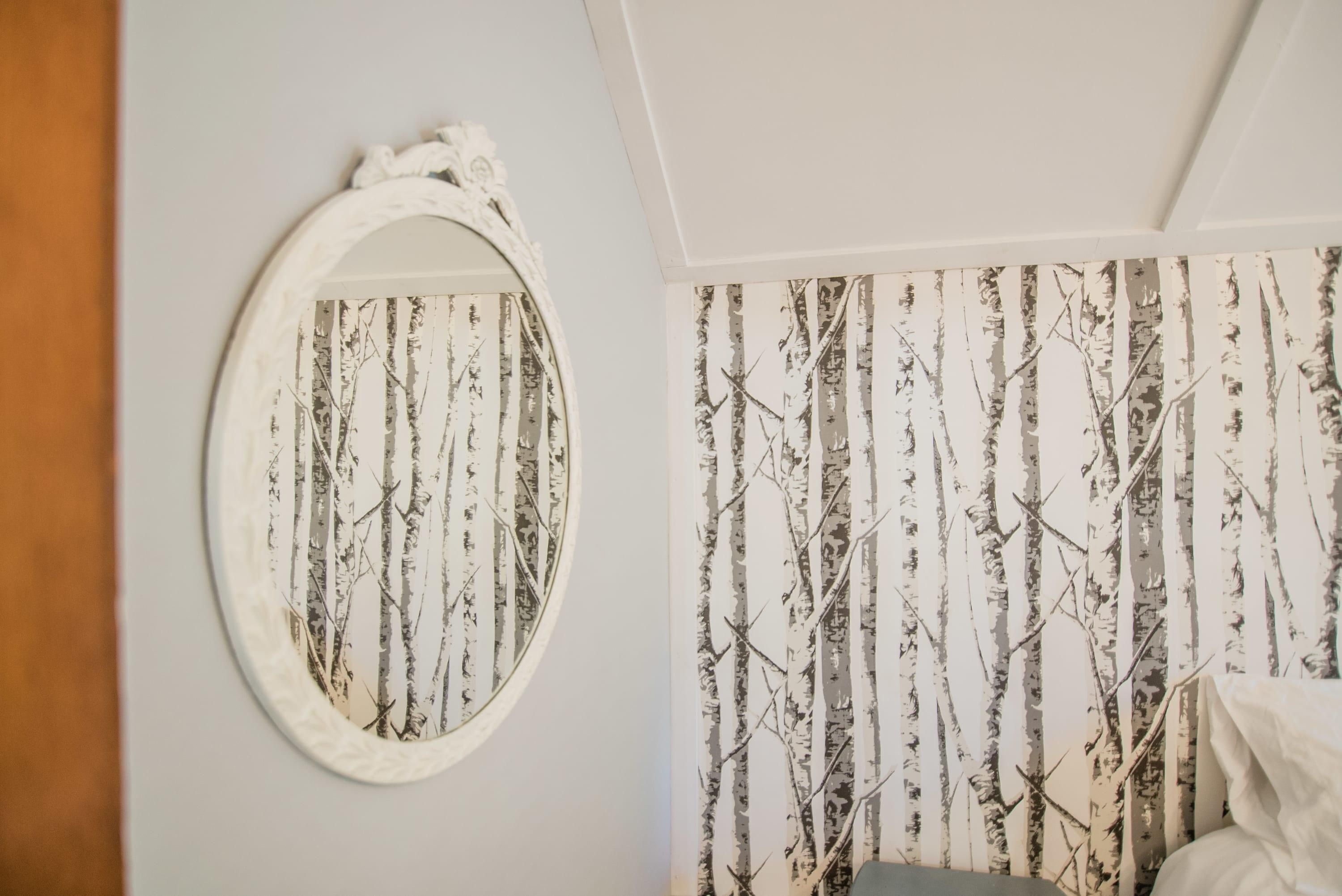A room with a white wall, a large oval mirror, and a wallpaper with vertical tree patterns.
