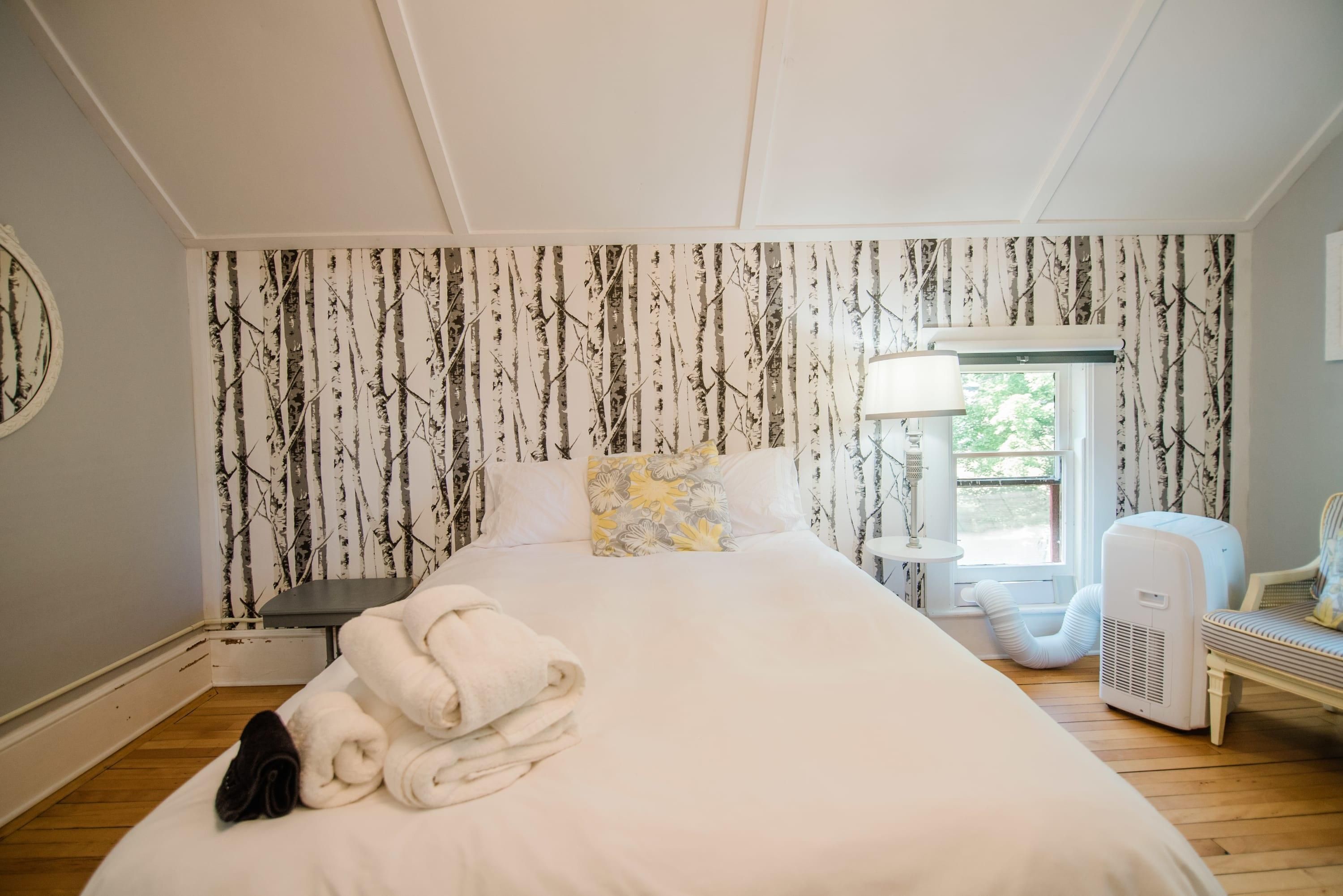 Quarto de hotel com uma cama grande, uma mesinha de cabeceira, uma luminária, uma janela com vista e uma parede com padrão de árvore.