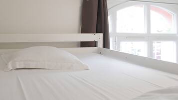 Ein Zimmer mit einem Einzelbett, einem weißen Kopfende, einem weißen Kissen und einem Fenster mit Vorhängen.