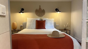 Een net opgemaakt bed met een rode deken, witte kussens en een strohoed erop. Aan beide zijden van het bed hangen twee wandlampen.