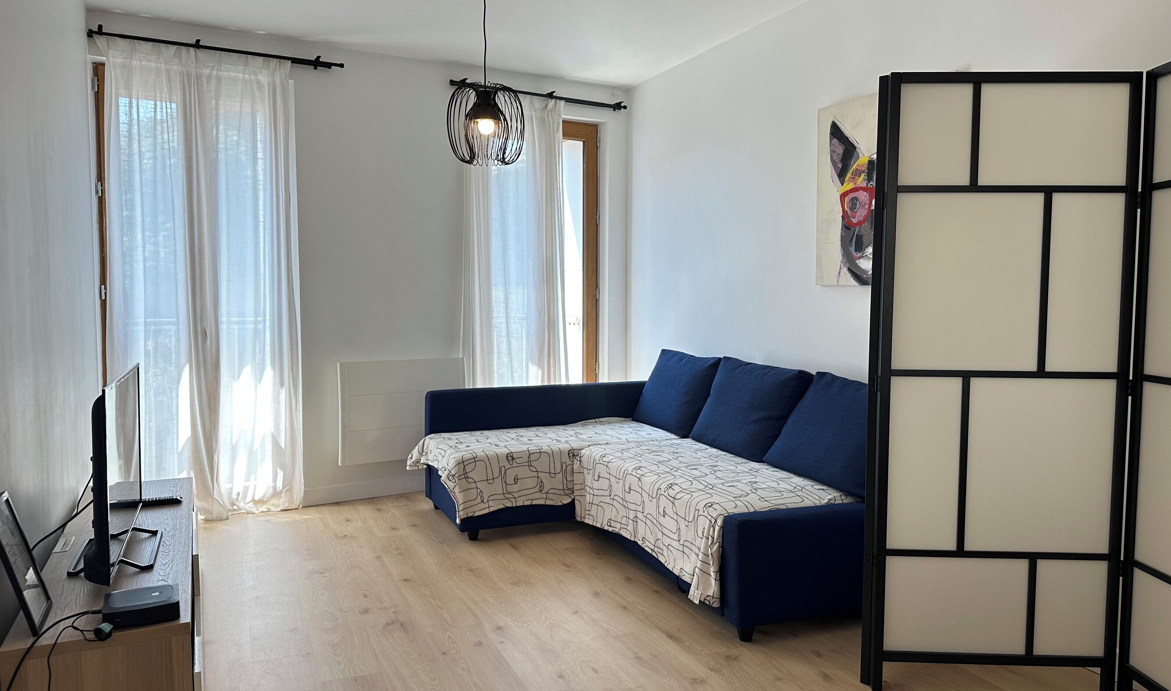 Une pièce moderne avec un canapé aux lignes courbes, un bureau debout équipé d’un ordinateur et une grande fenêtre ornée de rideaux légers.