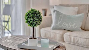 Una pequeña planta en maceta sobre una bandeja con una vela, sobre una mesa redonda de madera, ubicada en una sala de estar con un sofá y un cojín con la palabra 'love'.