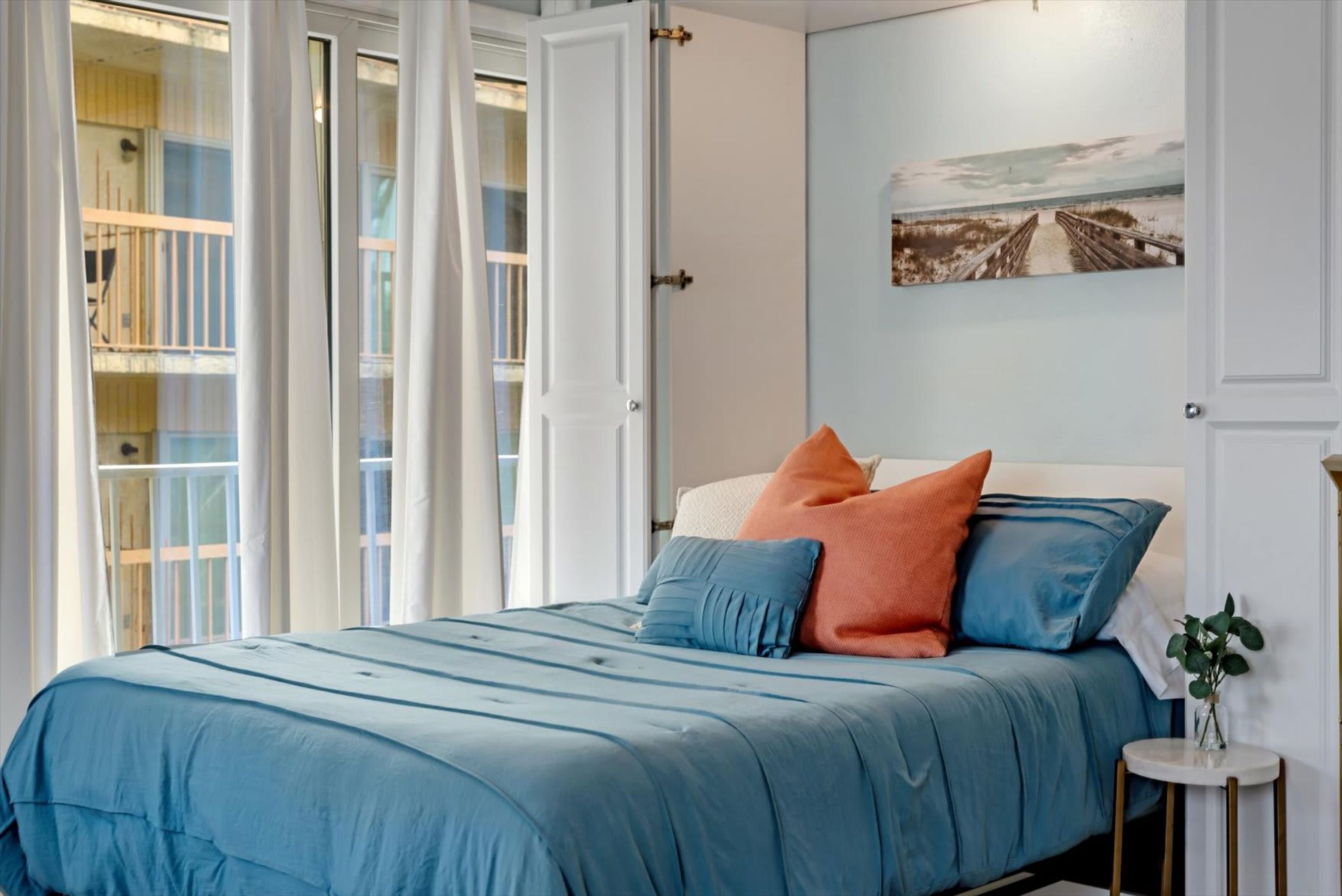 Un dormitorio con cama, almohadas, una mesita de noche, un cuadro en la pared y vistas a un balcón.