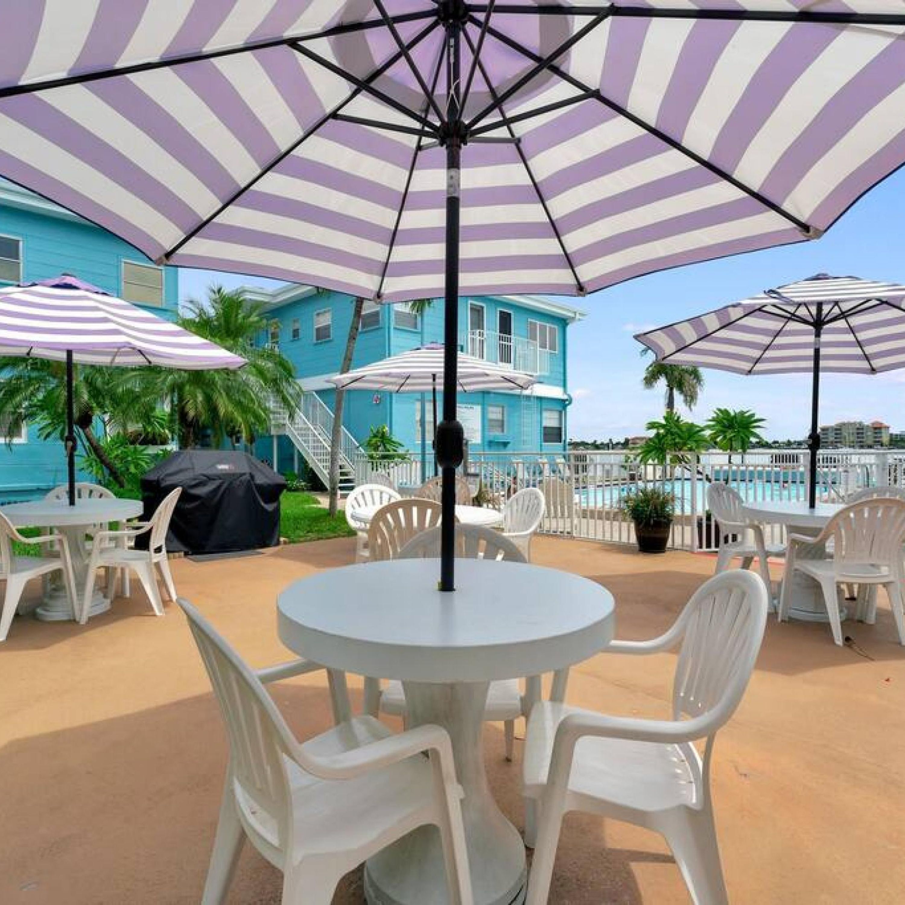 Un espace patio avec des chaises et des tables en plastique blanc, des parasols à rayures et une piscine en arrière-plan.