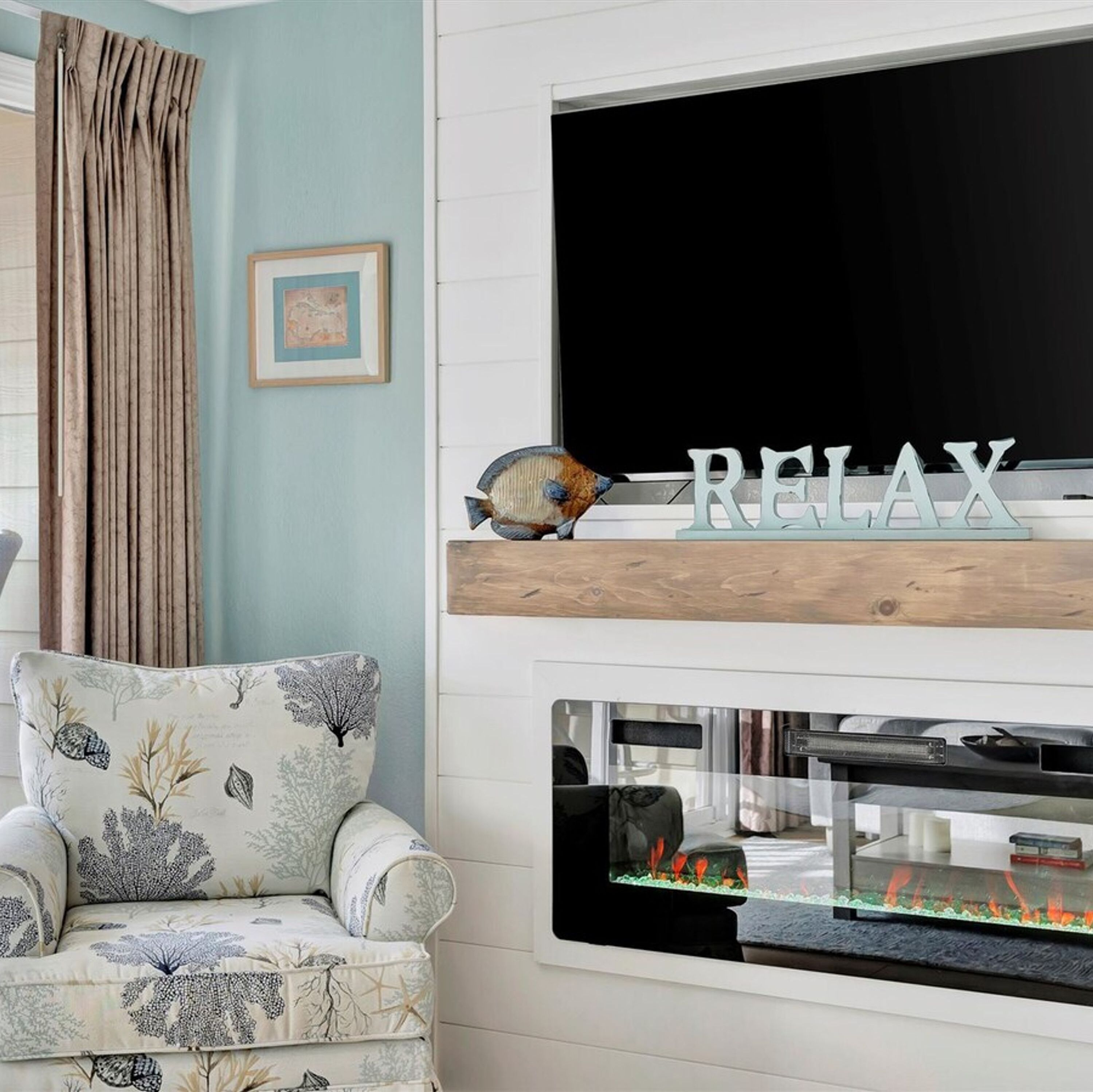 A living room with a TV mounted on a white wall, a fish tank below the TV, a chair with a nature-themed cushion, and a 'RELAX' sign on a wooden shelf.