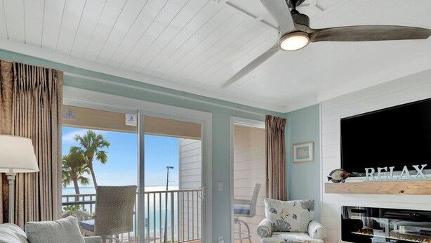 A living room with a sofa, a coffee table, a TV mounted on the wall, a ceiling fan, and a view of the ocean through the sliding glass doors. - Oceans Pointe Nantucket Inspired Condo on the bay (Tampa)