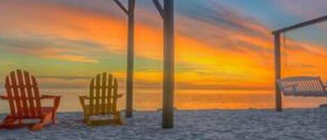 A beach scene with two Adirondack chairs and a wooden structure, likely a gazebo, under a sunset sky.