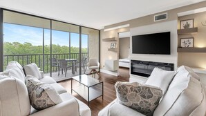 A modern living room with a large TV, comfortable seating, and a balcony with a view of greenery.