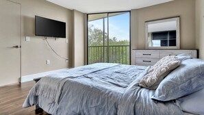 A bedroom with a large bed, a flat-screen TV mounted on the wall, a dresser, and a window with a view of greenery.