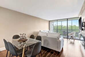 Sala moderna com mesa de vidro, sofás cinza e varanda com vista para o verde.