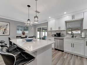 A modern kitchen with white cabinets, a central island, black bar stools, pendant lighting, and a view to the outside. - 4b 2b w Hot Tub BBQ Firepit 9mins to Beach (Largo)