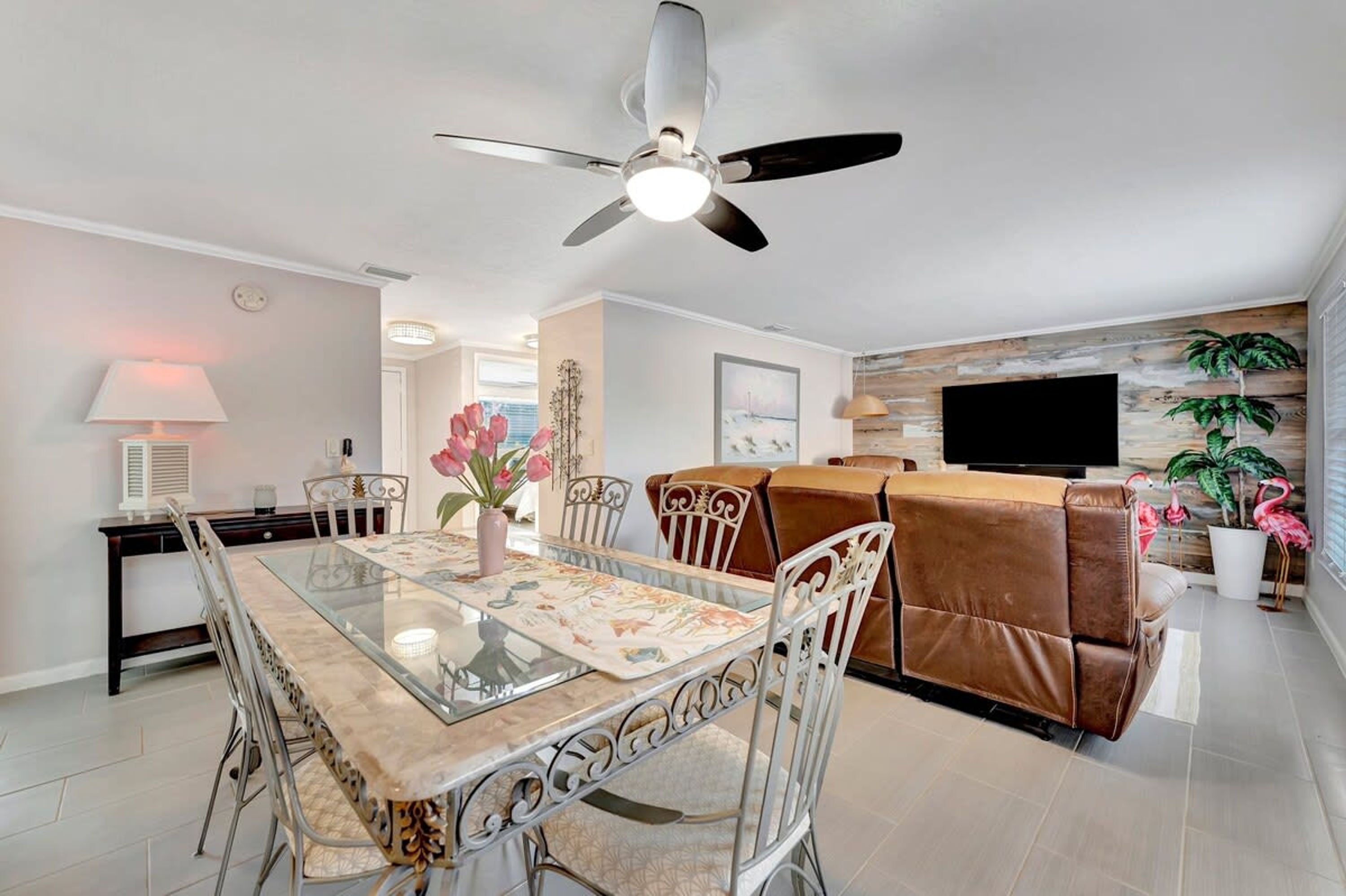A modern living room with a glass dining table, a brown leather sofa, a ceiling fan, and a flat-screen TV mounted on the wall.
