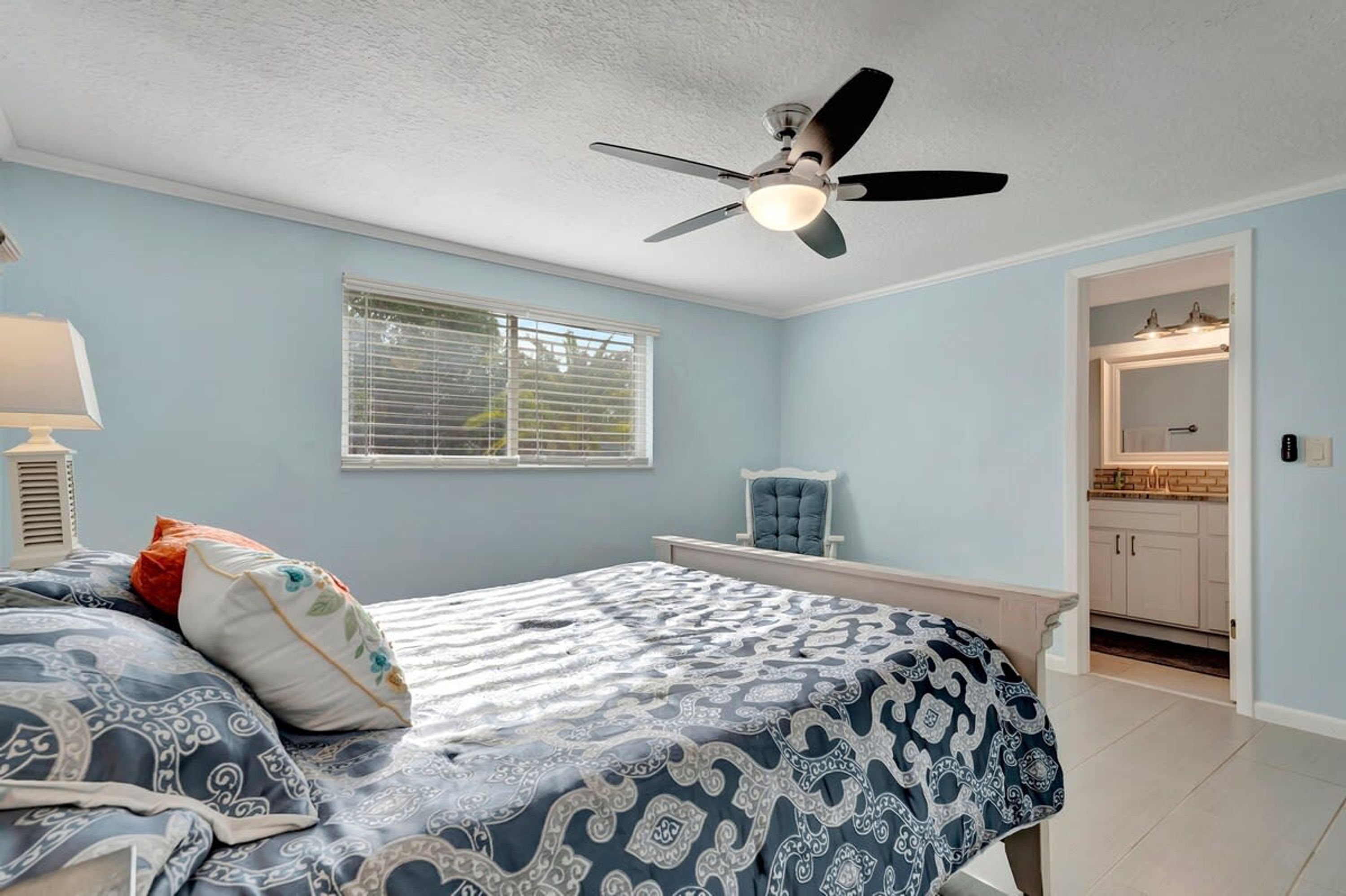 A bedroom with a bed, a ceiling fan, a window with blinds, a bedside table, and a view into the kitchen.