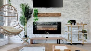 Un salon moderne avec un téléviseur à écran plat fixé à un mur en pierre, une cheminée en dessous, un fauteuil suspendu et une table basse en bois sur laquelle sont posés des livres et des télécommandes.