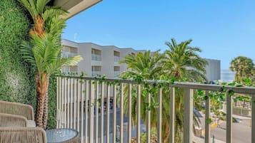 A balcony with wicker furniture, a palm tree, and a view of a hotel building and street.
