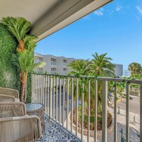 En balkon med fletningsmøbler, en palme og udsigt til en hotelbygning og gade.