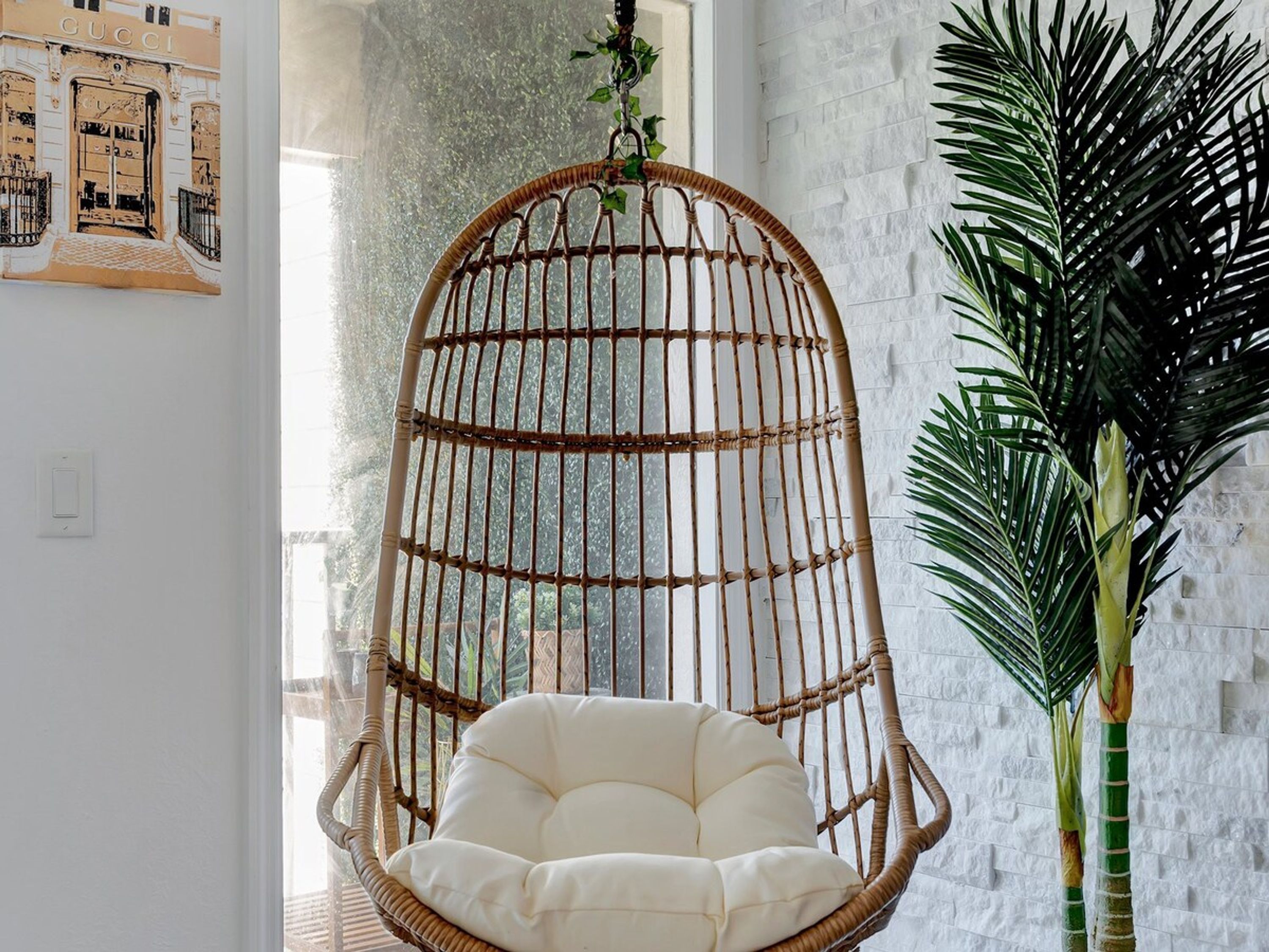 Una silla colgante de mimbre con un cojín, una planta en maceta y una pared con un cuadro enmarcado.