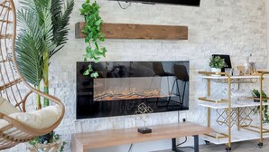 Un salon avec un mur en pierre, un téléviseur à écran plat fixé au-dessus d’une cheminée, un banc en bois, un fauteuil en osier et un chariot doré contenant de la verrerie.