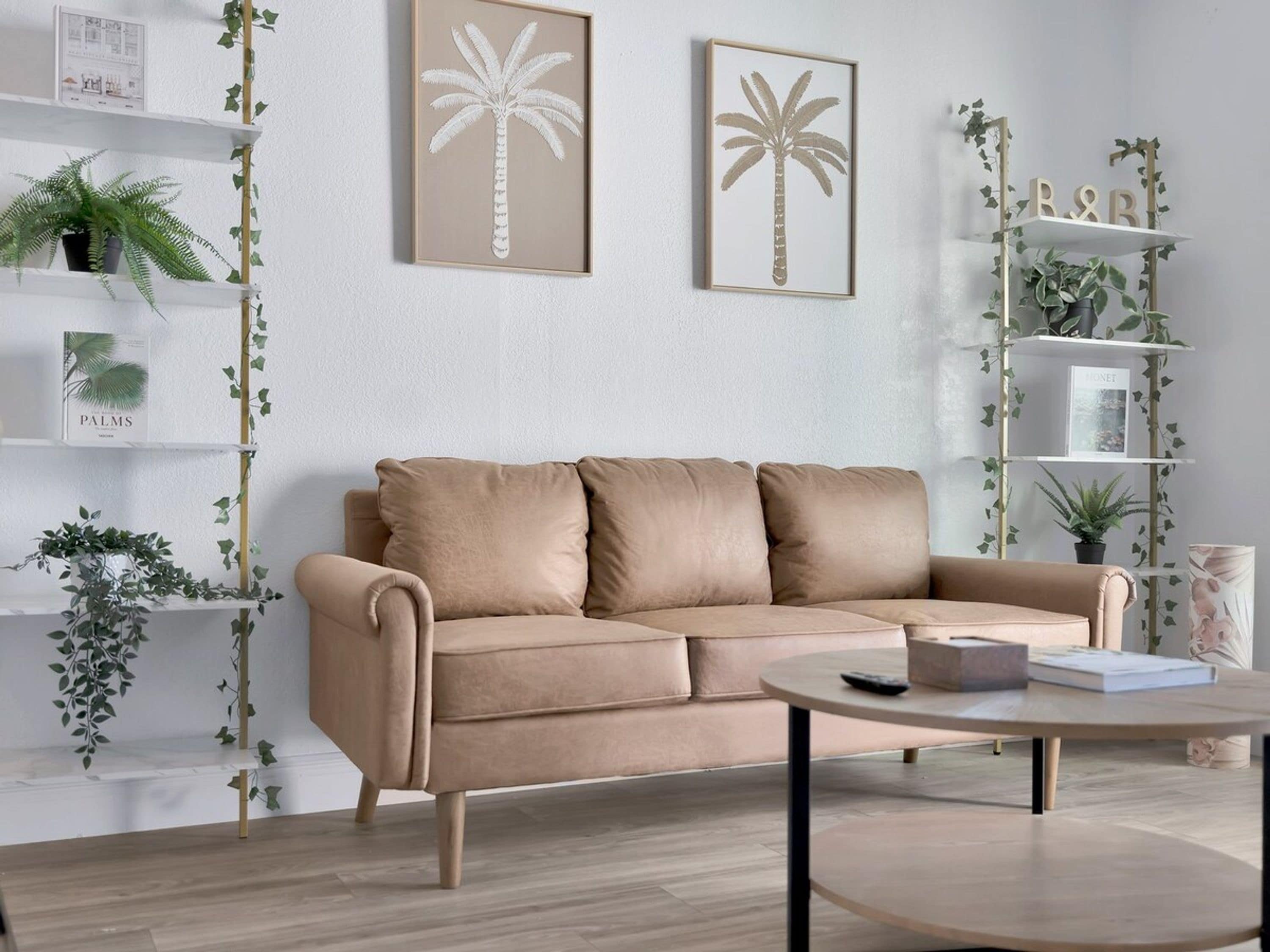 En moderne stue med en beige sofa, et rundt sofabord og en reol med planter.