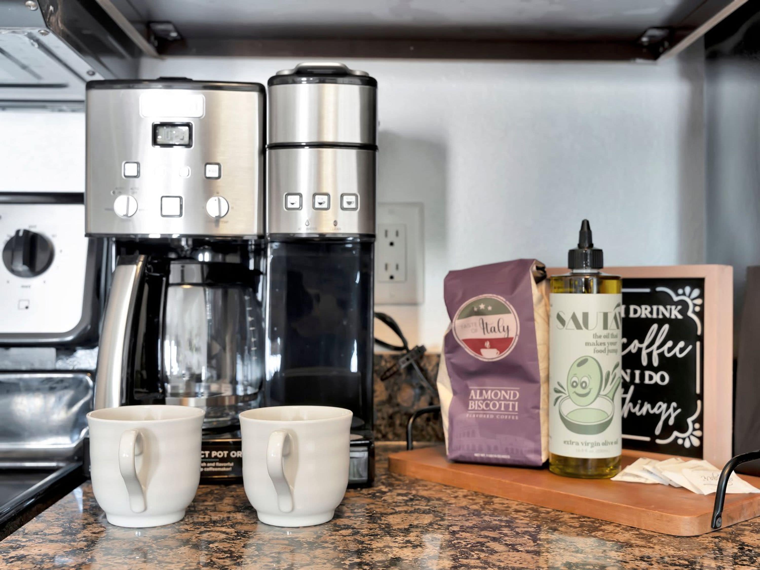 Una encimera de cocina con una cafetera, dos tazas blancas, un paquete de biscotti de almendras, una botella de aceite de oliva virgen extra, y un letrero que dice 'DRINK COFFEE IN DO'.