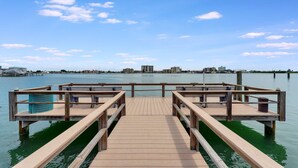 Eine Holzstege zu einem Anlegesteg mit Bänken, der sich über ruhiges Wasser mit blauem Himmel und weit entfernten Gebäuden erstreckt.