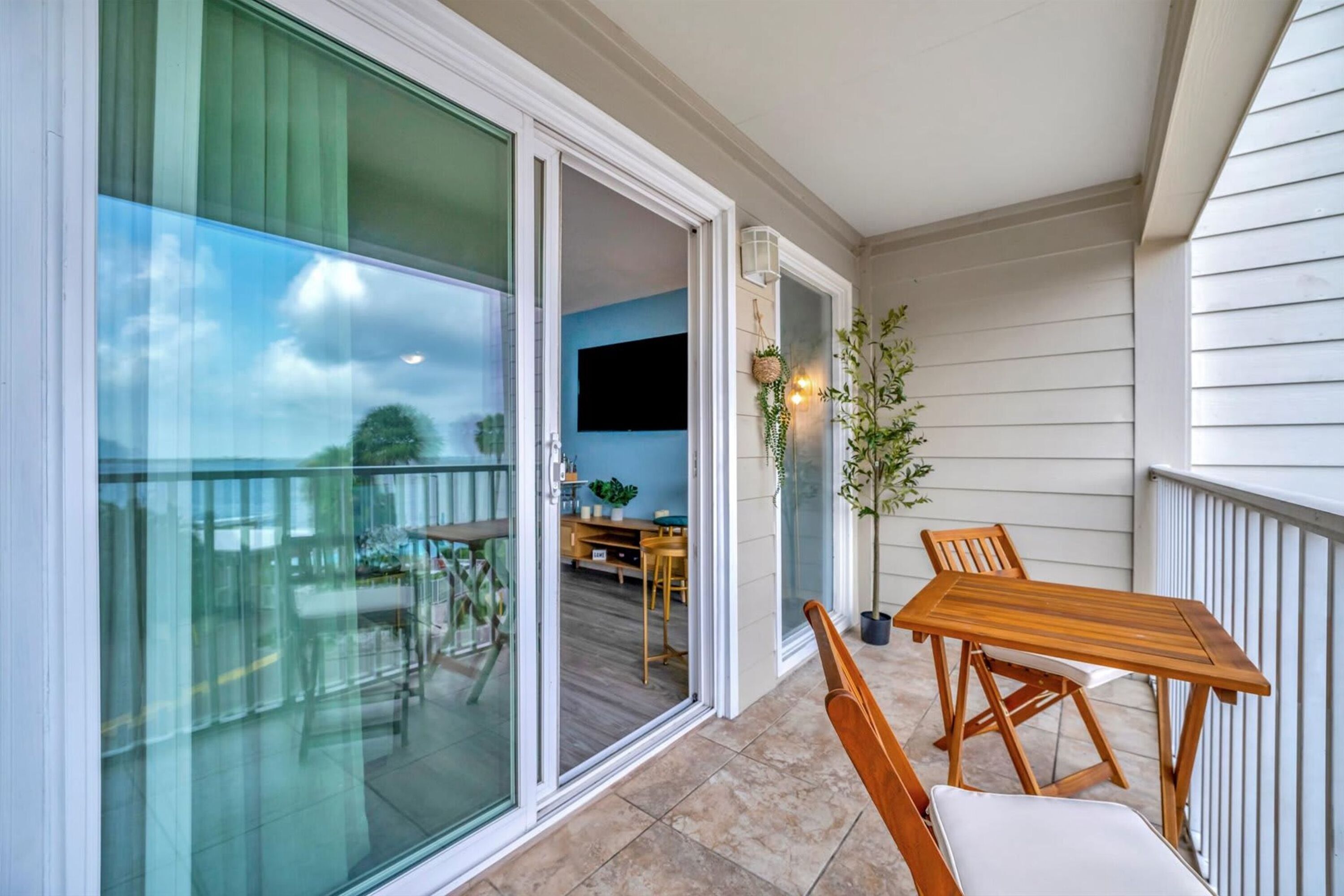 An outdoor balcony with a wooden table and chairs, a potted plant, and a view of the interior dining area.
