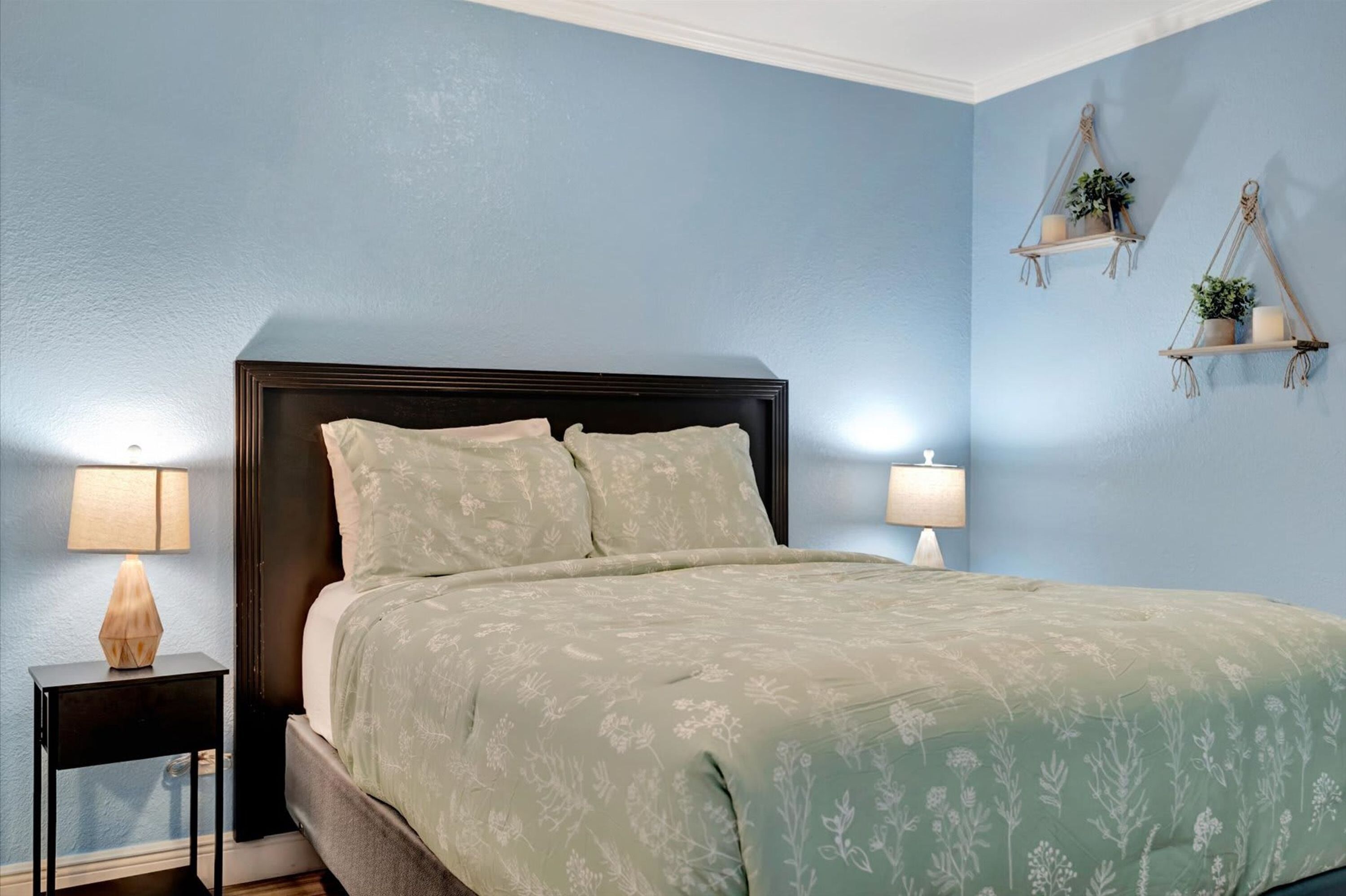 Un dormitorio con cama, dos lámparas de noche, una mesita y estantes en la pared con plantas.
