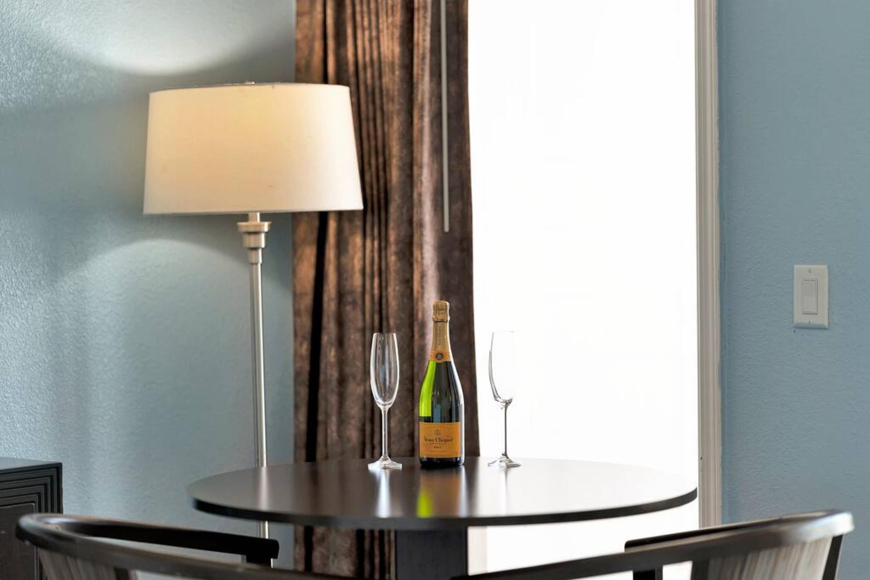 A round table with a white table lamp, a bottle of champagne, and two champagne flutes.