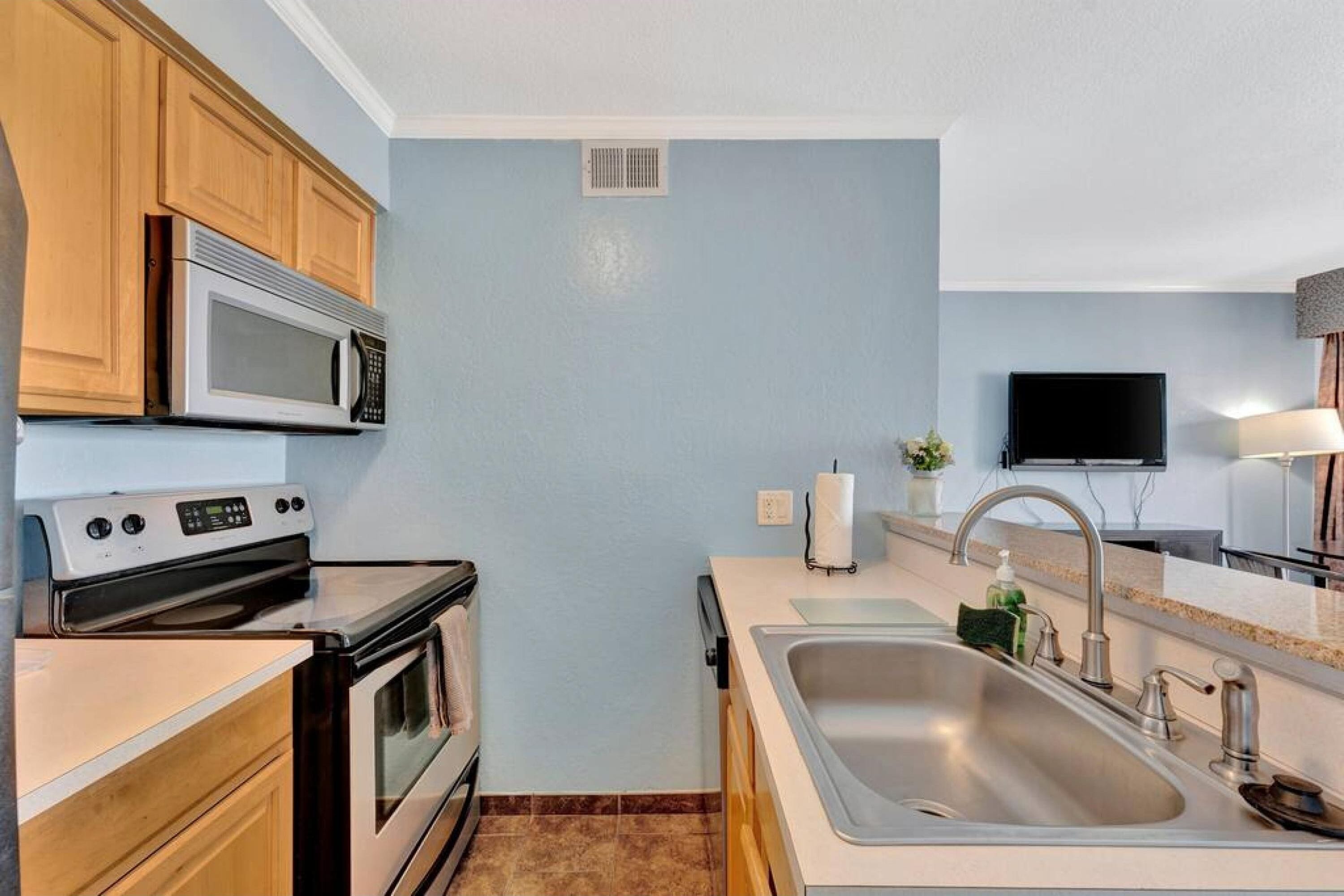A compact kitchen with wooden cabinets, a microwave, stove, sink, and a flat-screen TV mounted on the wall.