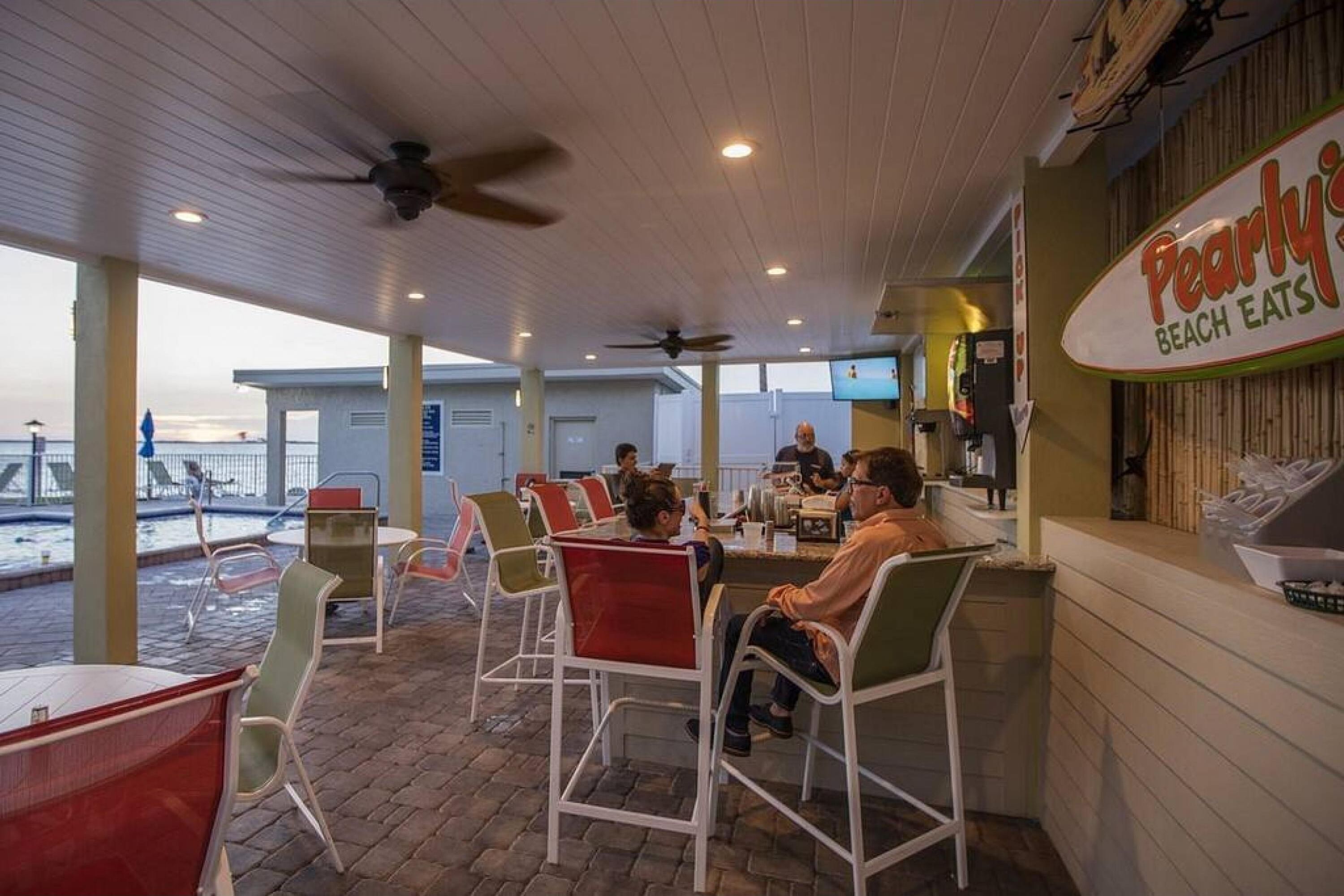 Un espace extérieur aménagé avec des tables et des chaises, une enseigne indiquant « Pearly’s Beach Eats », et une vue sur la plage en arrière-plan.