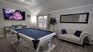Een woonkamer met een pooltafel, een televisie, een bank en een groot raam.