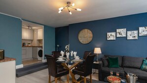 A modern living room with a dark blue accent wall, a round coffee table, a sofa, and a dining area with a glass table and chairs. - Short Walk to I360 and Beach 5 Condo With Patio (Brighton)