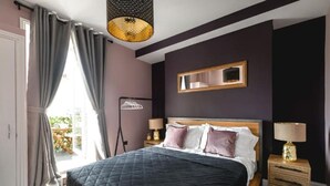 A bedroom with a bed, a bedside table, a lamp, a mirror, and a window with curtains. - Designer Retreat 5 Location Balcony Parking (Hove)