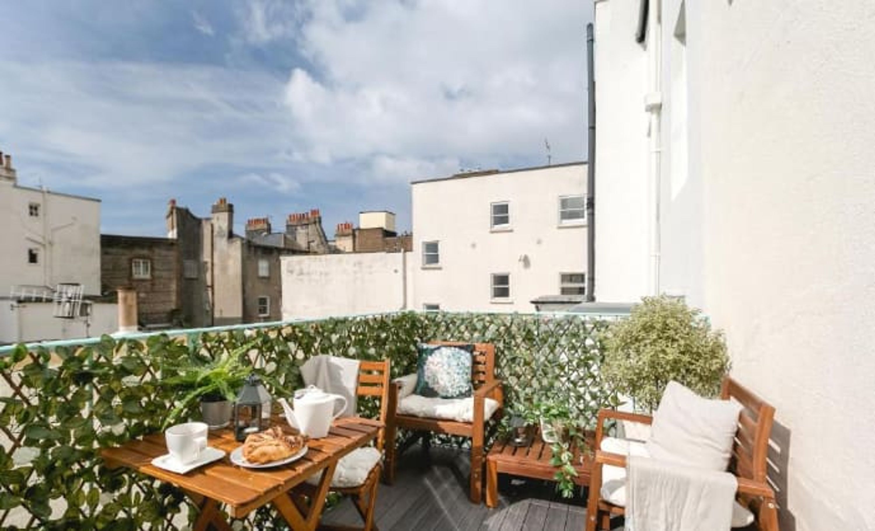 A rooftop terrace with a wooden table set for tea, surrounded by chairs and potted plants.