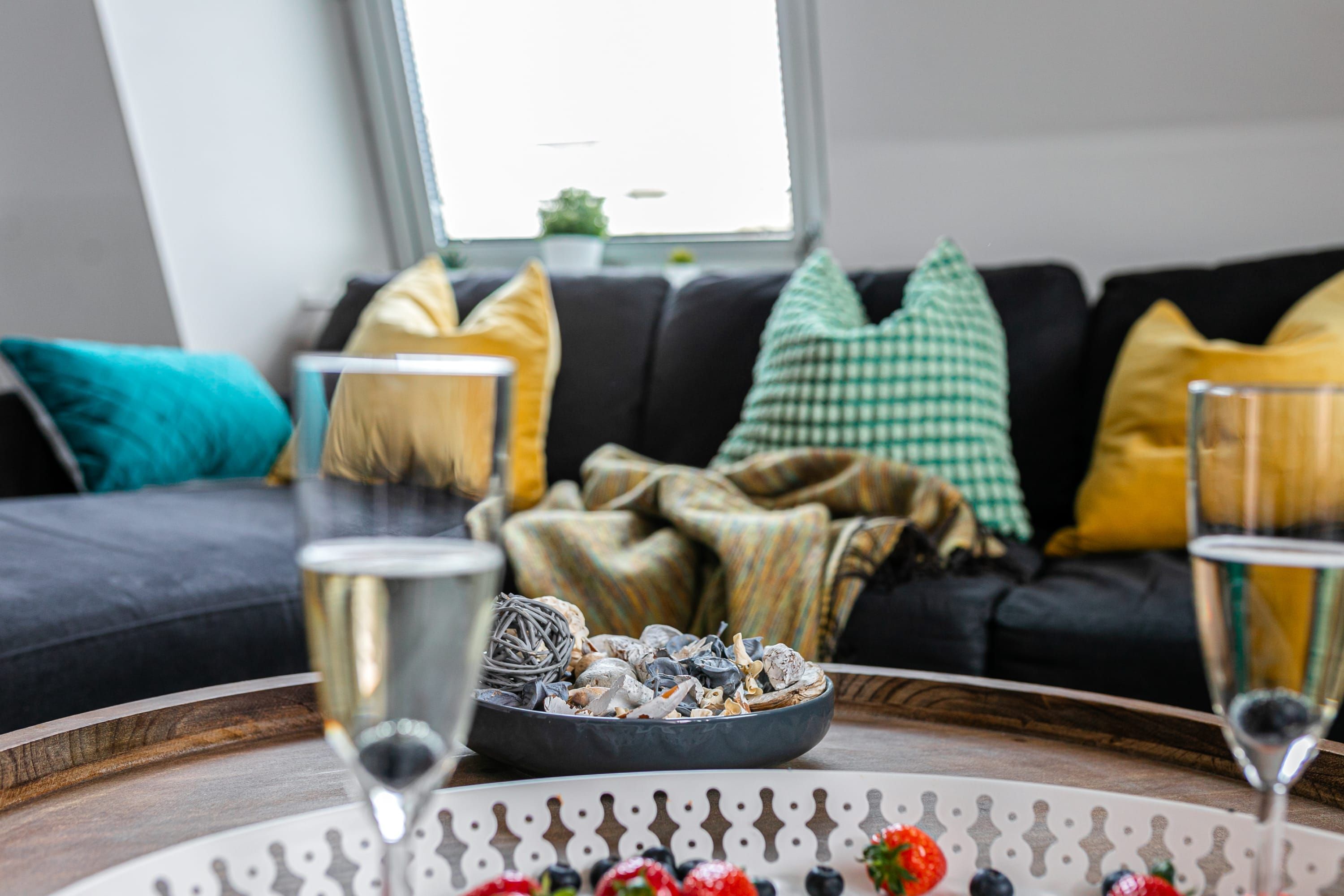Una sala de estar con un sofá negro, cojines coloridos, un cuenco de aperitivos y dos copas de vino sobre una mesa de madera.