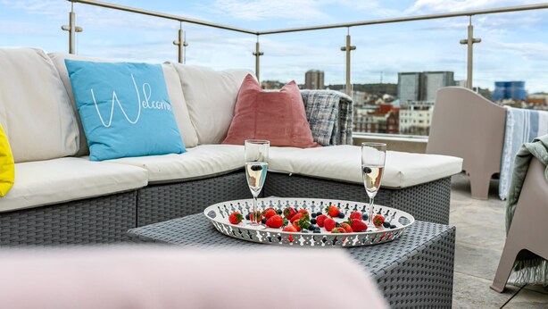 A rooftop terrace with a wicker sofa, a tray of strawberries and champagne glasses, and a city view in the background. - The Lanes Gem Penthouse Rooftop Terrace Sea View (Brighton)
