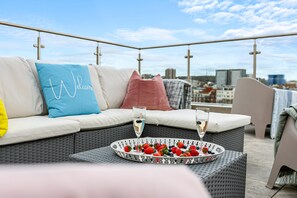 A rooftop terrace with a wicker sofa, a tray of strawberries and champagne glasses, and a city view in the background.