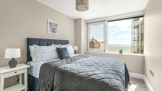 A bedroom with a large bed, a bedside table with a lamp, a window with a view of the sea, and a geometric pendant light. - Brighton Lanes Sea View 2BR AP (Brighton)