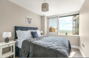 Un dormitorio con una cama grande, una mesita de noche con lámpara, una ventana con vista al mar y una lámpara colgante de diseño geométrico.