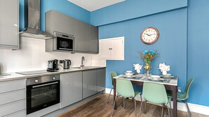 A modern kitchen with stainless steel appliances, a dining area with a wooden table and green chairs, and a blue accent wall. - The Lanes Getaway Moments Away From Pier Beach (Brighton)
