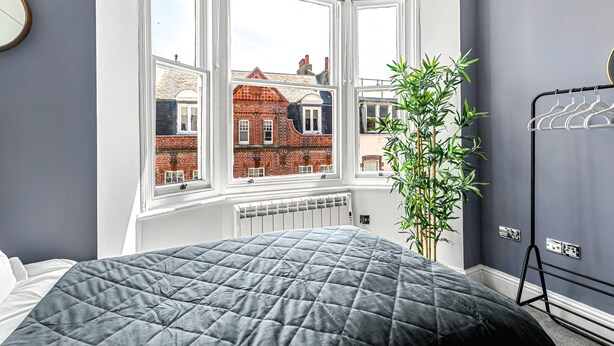 A bedroom with a large window, a radiator, a bed with a grey quilt, and a plant. - The Lanes in Brighton by Creatick (Brighton)