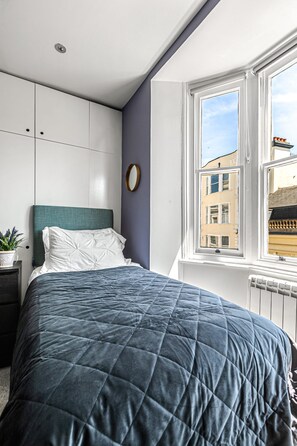 Un dormitorio con cama, mesita de noche, planta en maceta y ventana con vista a edificios.