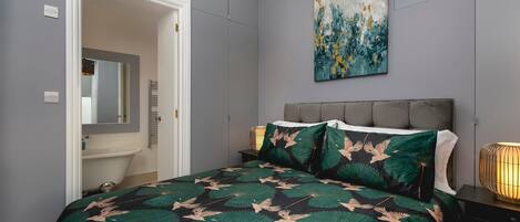 A bedroom with a bed, a nightstand, a wall-mounted lamp, a painting, and a bathroom visible through an open door.