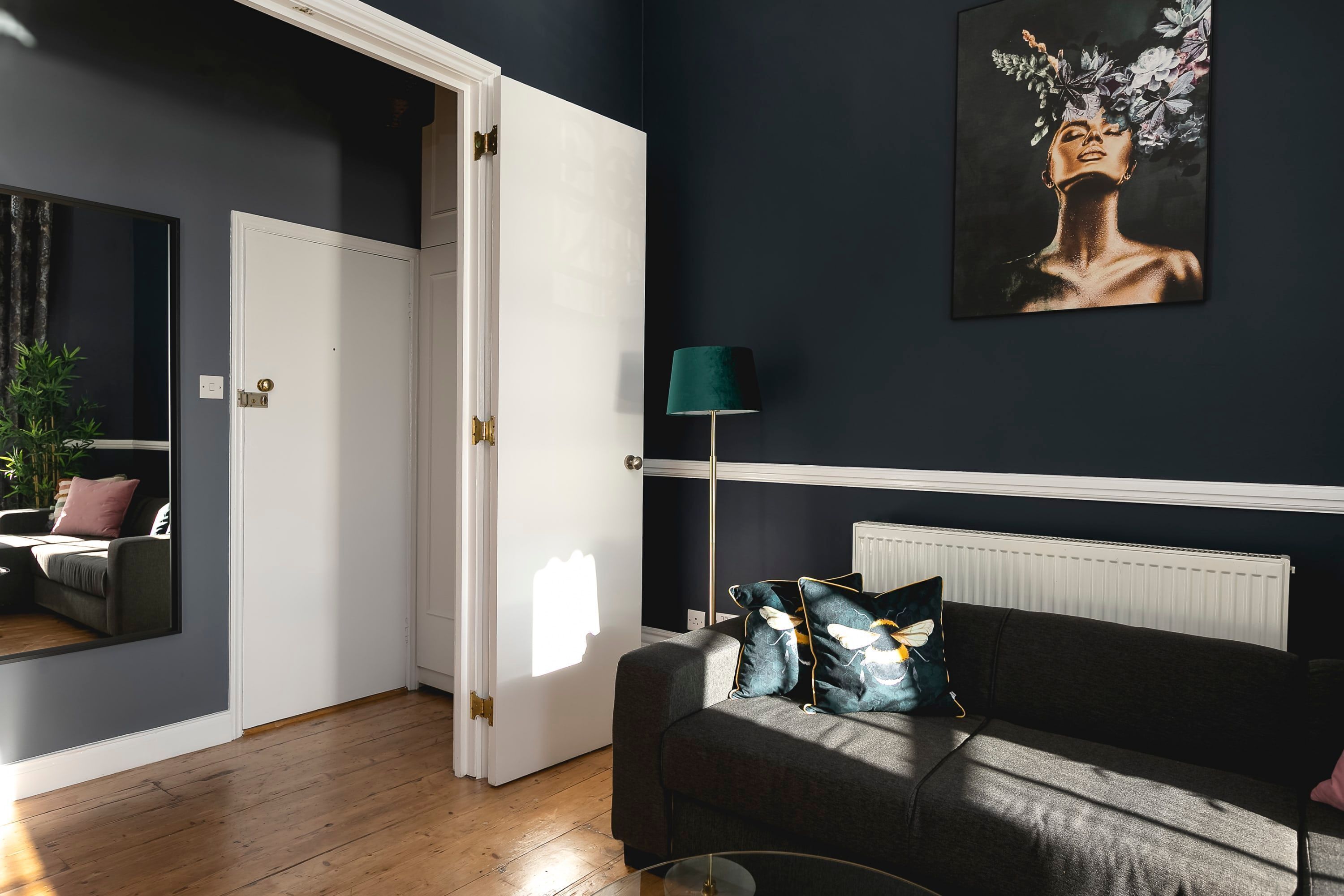Un salon moderne avec un mur bleu foncé, une porte blanche, un canapé gris agrémenté de coussins décoratifs, un lampadaire et une œuvre d’art encadrée.